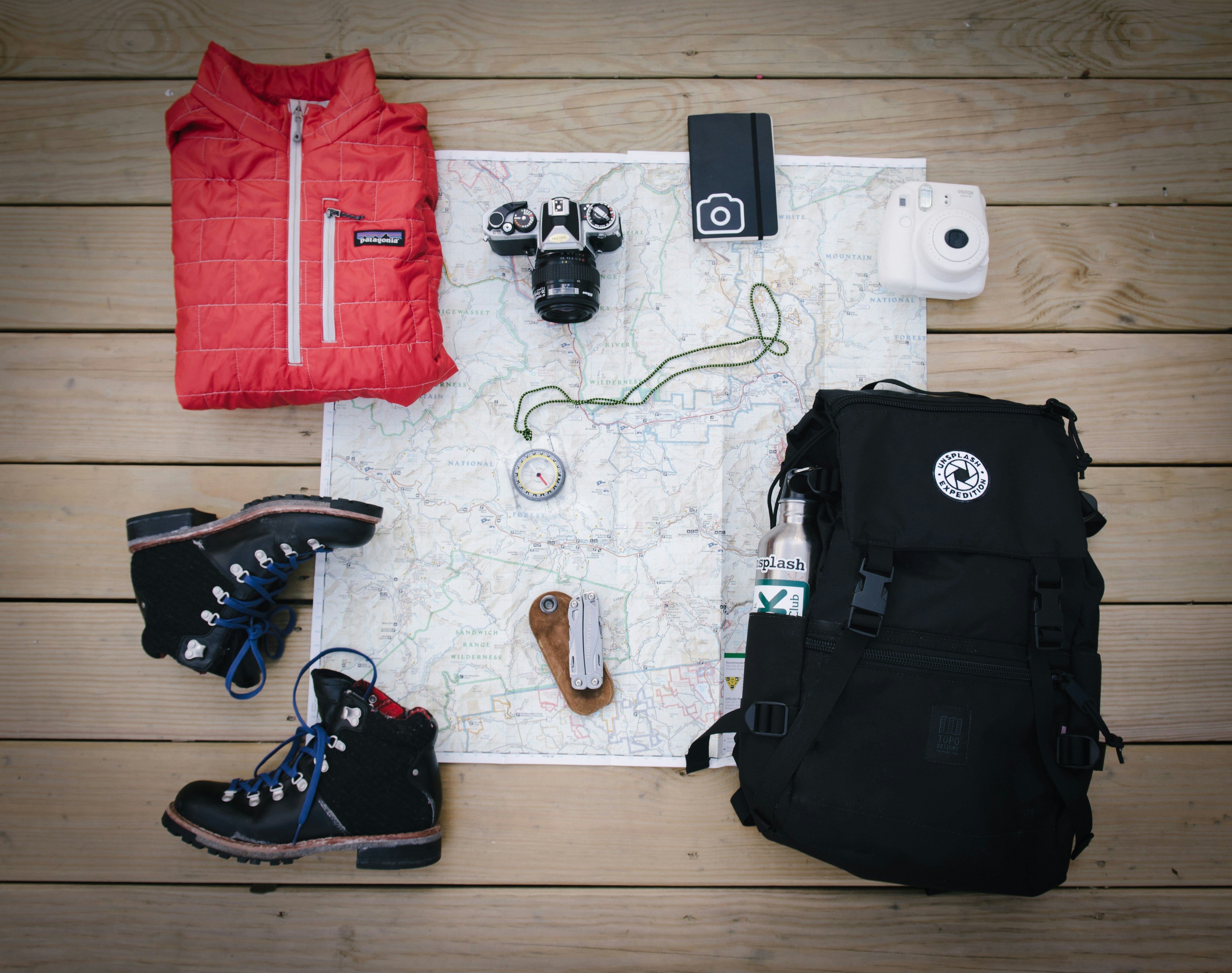 Travel and hiking essentials laid out on wooden planks, including a map, camera, boots, backpack, and a red vest.