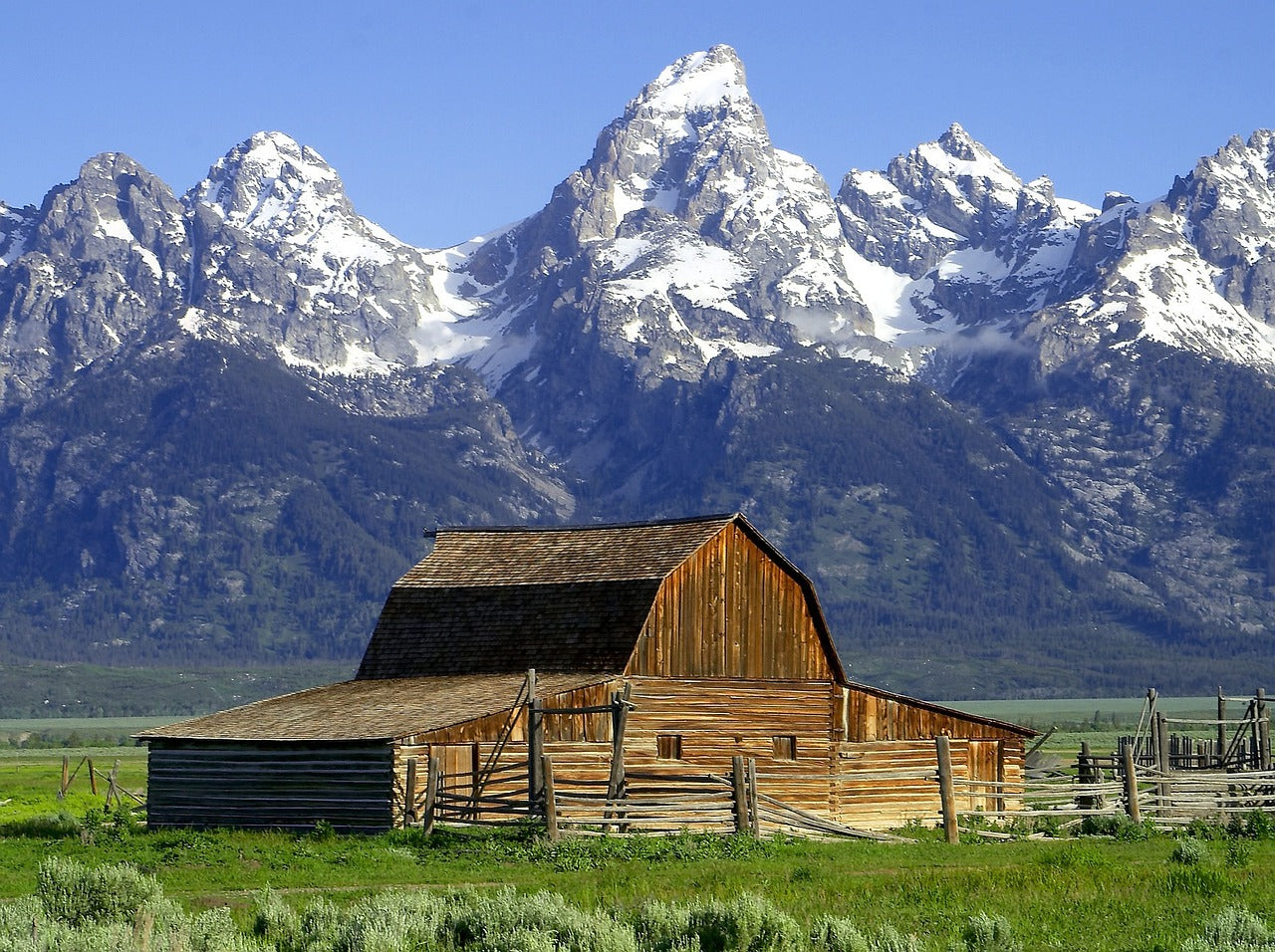 A wooden farm in a grassy field with a backdrop of snow-covered mountains